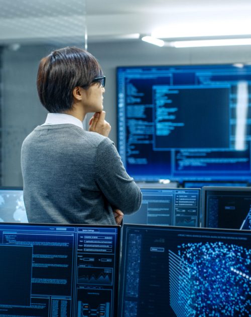 In the System Control Room Technical Operator Stands and Monitors Various Activities Showing on Multiple Displays with Graphics. Administrator Monitors Work of  Artificial Intelligence, Big Data Mining, Neural Network, Surveillance Project.